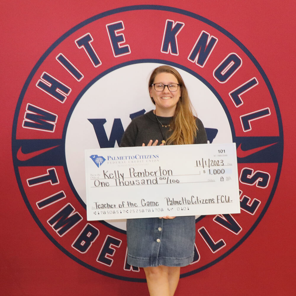 A smiling woman holding a $1,000 check for 'Teacher of the Game' in front of a White Knoll logo.
